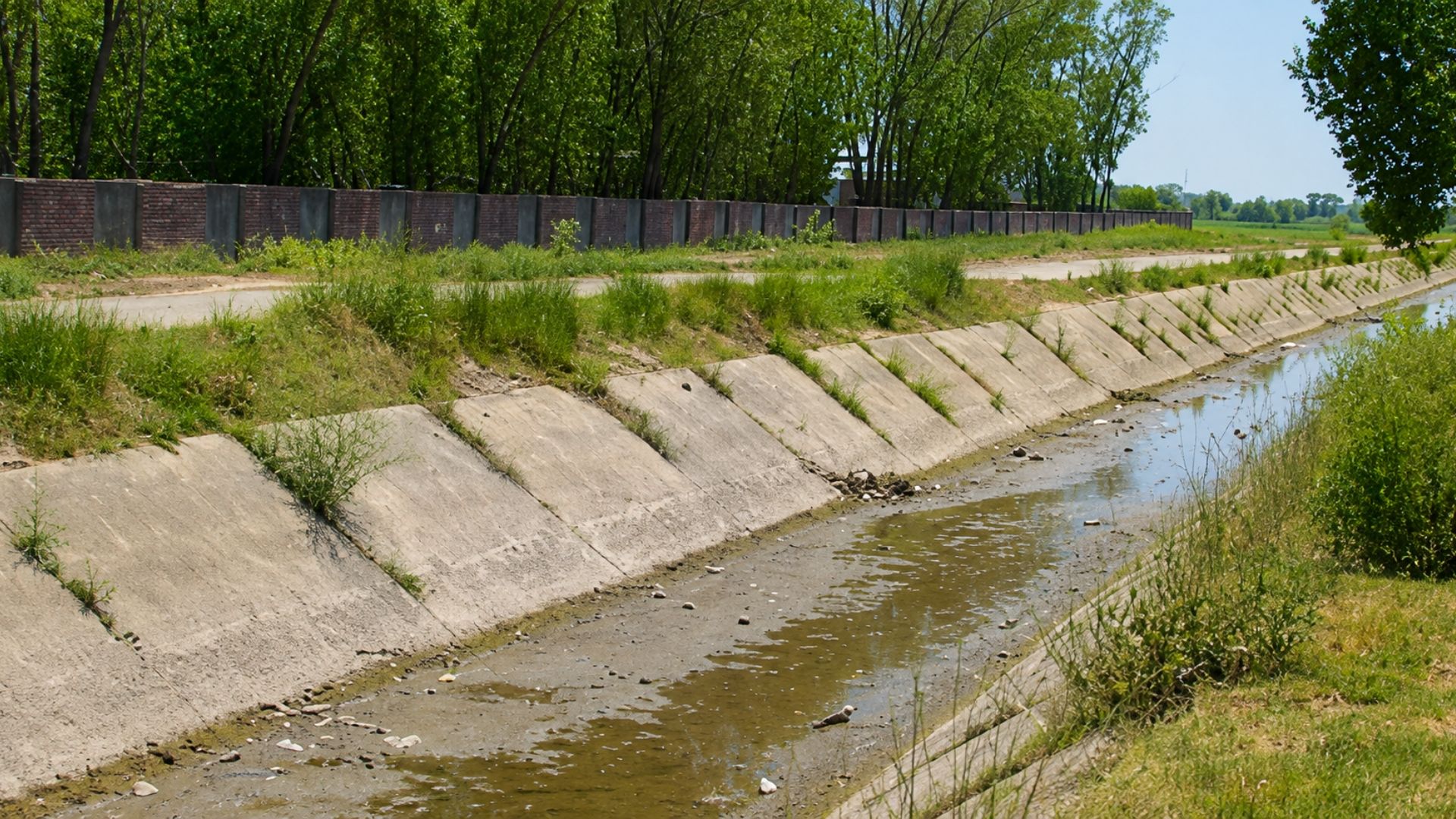 Concrete drainage canal with shallow water beside a road and trees on a sunny day