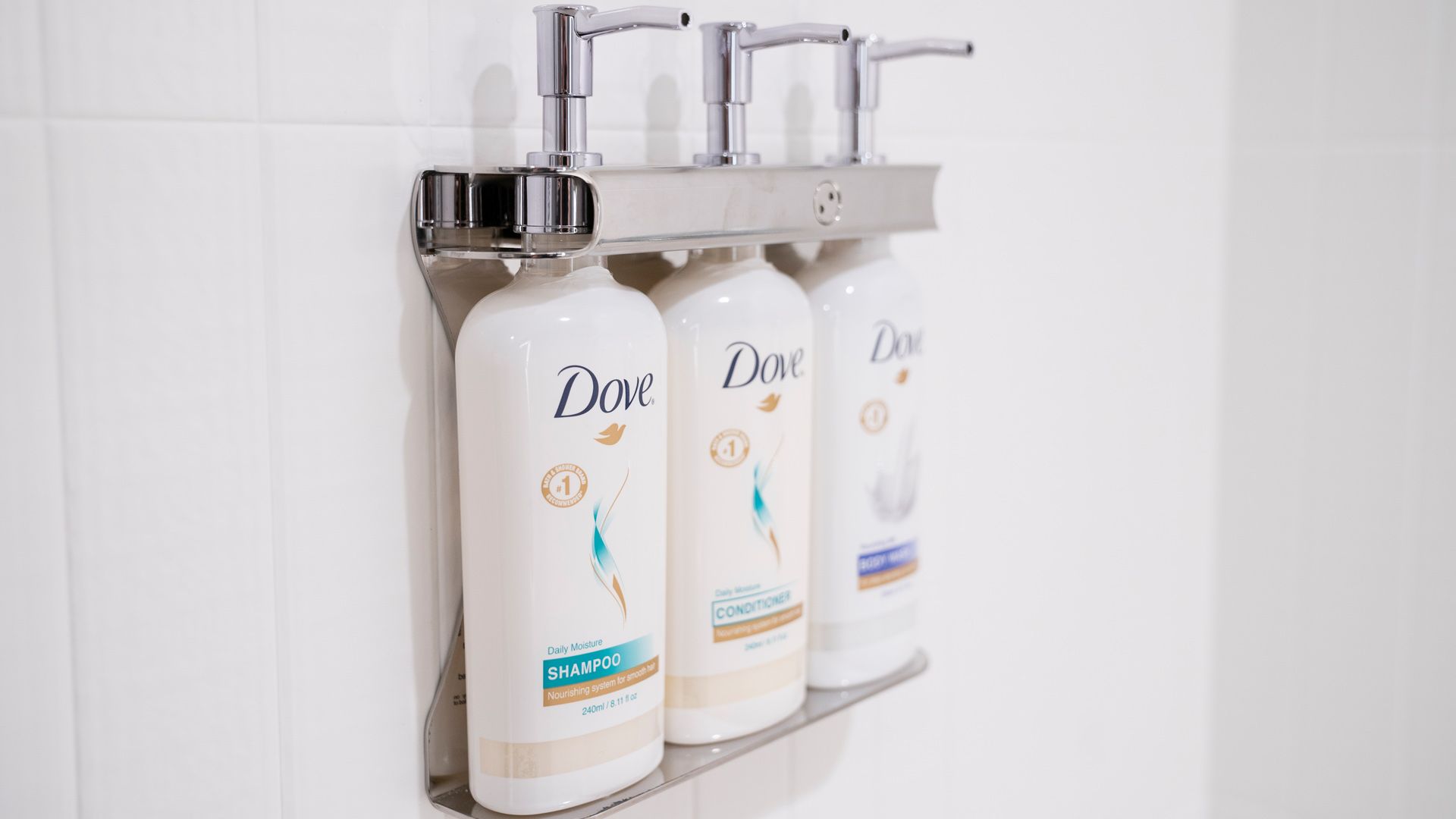 Bottles of Dove shampoo, conditioner and body wash in silver pump dispensers on a white tiled bathroom wall.