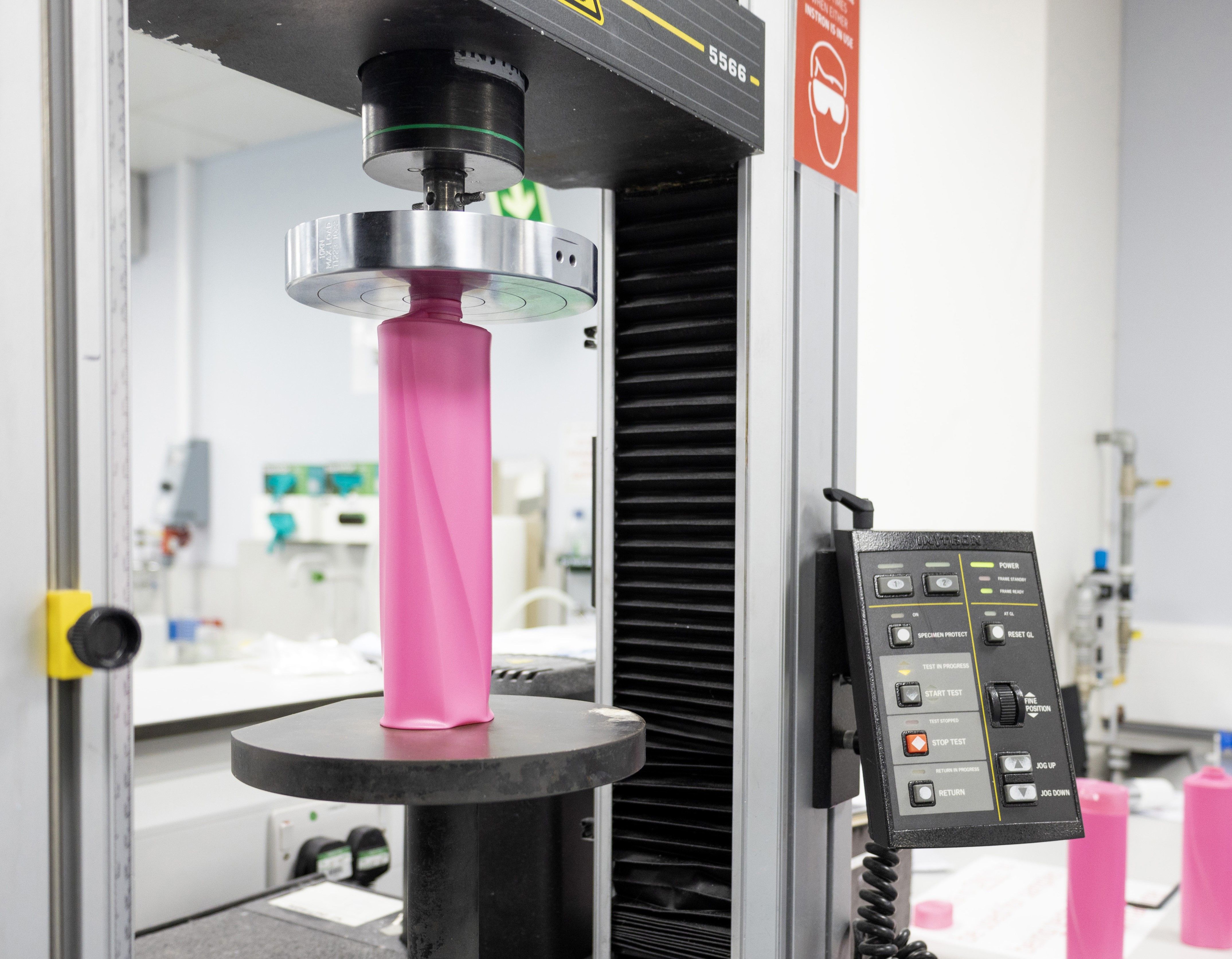 A close-up view of some lab testing equipment, showing a pink plastic bottle being compressed and tested in Unilever’s packaging R&D lab. The machine's control panel with various buttons is visible on the right.