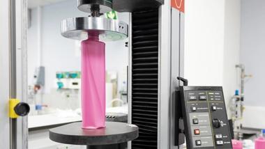 A close-up view of some lab testing equipment, showing a pink plastic bottle being compressed and tested in Unilever’s packaging R&D lab. The machine's control panel with various buttons is visible on the right.