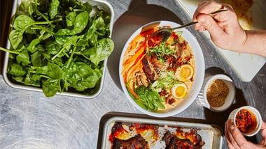 An image of dishes on a stainless-steel table, including a tray of green leaves and a ramen bowl over which red sauce is being drizzled.