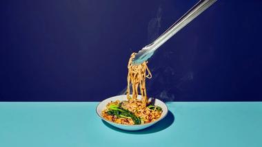 An image of Asian-inspired noodles being lifted from a white bowl  with tongs, shot against a split background of dark and light blue.