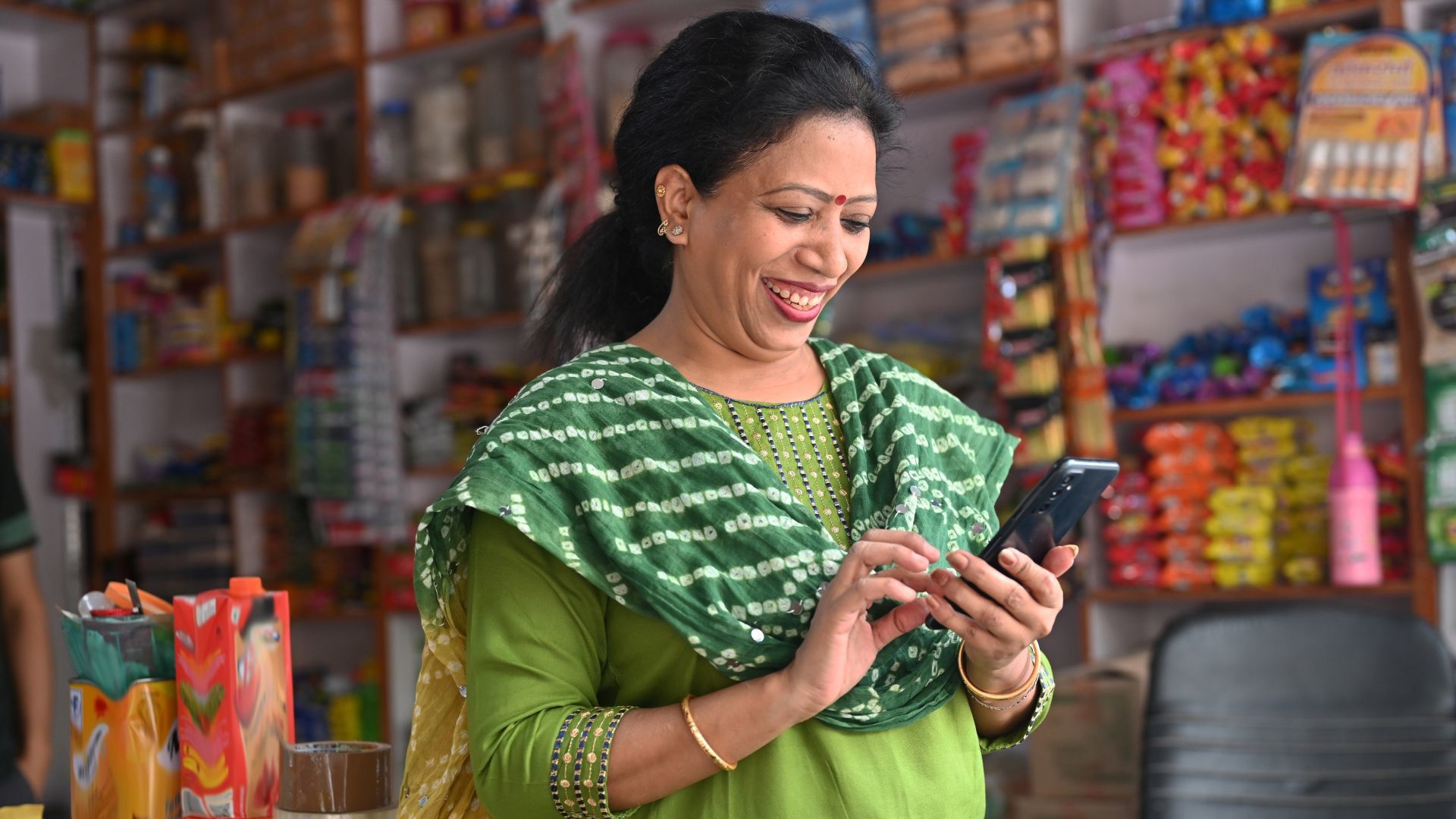 A woman in green traditional attire using a smartphone inside a colorful small shop with shelves of various products.