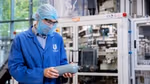 A person wearing a blue Unilever lab coat, hairnet, safety glasses and face mask examines a grey plastic bottle in a factory setting, with industrial equipment in the background.