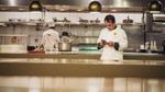 A chef consulting his phone in a professional kitchen while another chef works  in the background